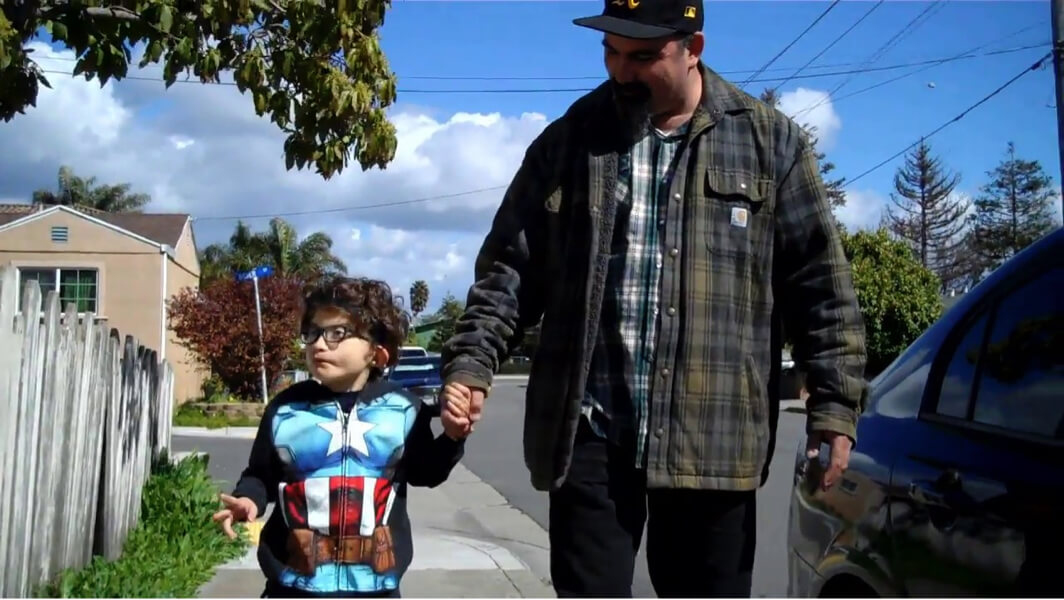 A dad holds his son's hand as they walk on the sidewalk in a neighborhood.
